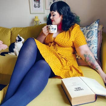 Person successful a yellowish polka-dot formal and navy bluish tights holding a cat-themed mug, sitting connected a sofa with a feline and an unfastened publication nearby