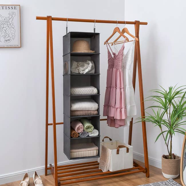 A clothing rack with shelves holding folded items and a pink dress hanging next to hats. A bag and shoes on the floor