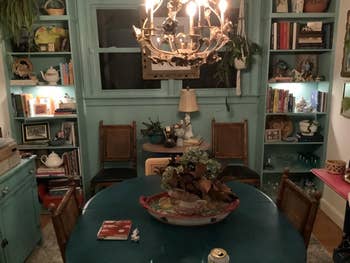 Dining country   with a chandelier, woody  chairs, and a decorative centerpiece connected  a circular  table. Books and decorative items capable    the shelves