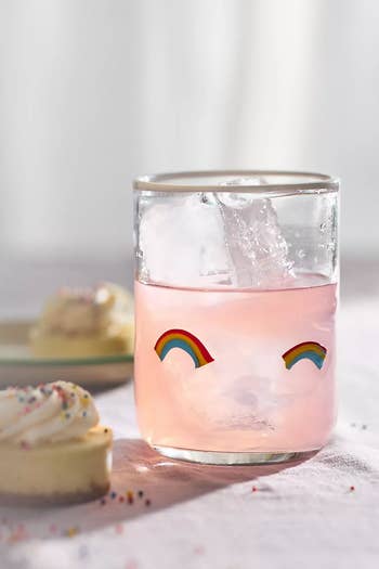 Glass with pink drink, ice, and rainbow designs on the front; a cupcake with sprinkles is in the background