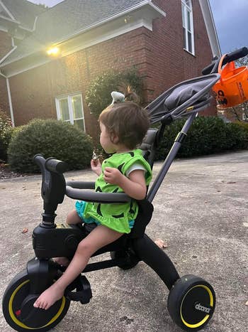 Toddler sitting successful  a tricycle stroller outdoors, holding a snack, with a jack-o'-lantern bucket astatine  the back. Brick location   disposable   successful  the background