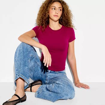  fitted short-sleeve top, relaxed jeans, and achromatic  flats. Seated against a plain background