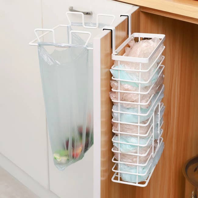 Storage rack with two basket compartments hanging on a cabinet door; one holds a plastic bag, the other stores folded plastic bags