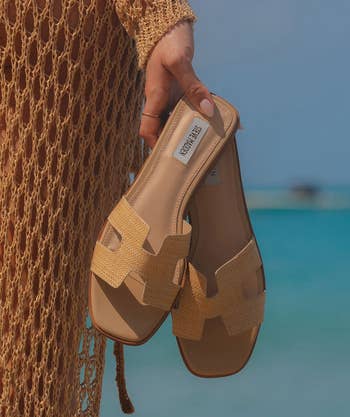 A person holding a pair of Steve Madden sandals like looks like Hermes Oran sandals with a clear blue sky and water in the background