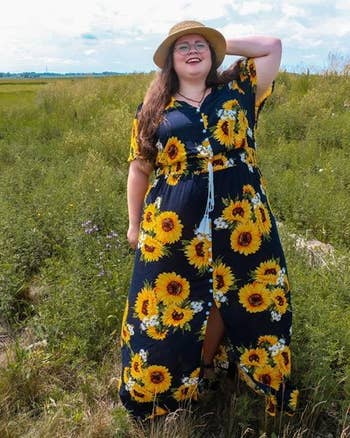 Reviewer in a sunflower print dress and hat stands in a field, posing with hand on hip