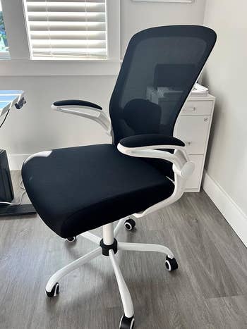 Office chair with mesh back, cushioned seat, armrests, and wheels. Positioned in a modern home office setting near a desk and cabinet