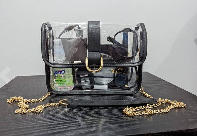 Clear purse with black seams and flap over clasp with a gold chain 