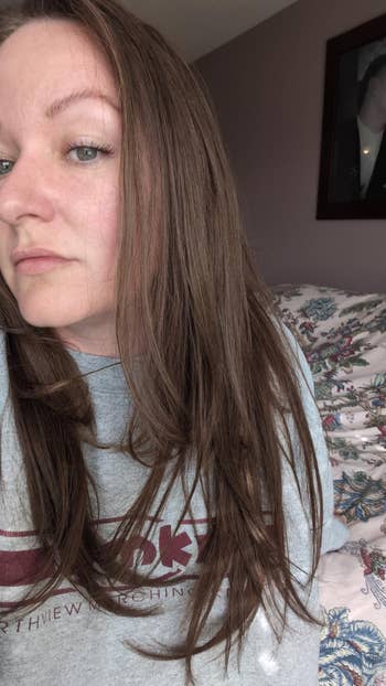 A person indoors with long hair, wearing a casual graphic sweatshirt, sitting on a bed with a patterned blanket