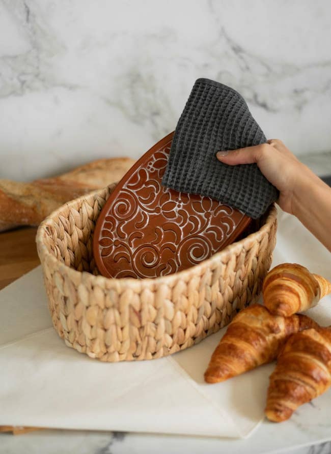 A manus placing a patterned clay breadstuff warmer into a woven handbasket with croissants nearby