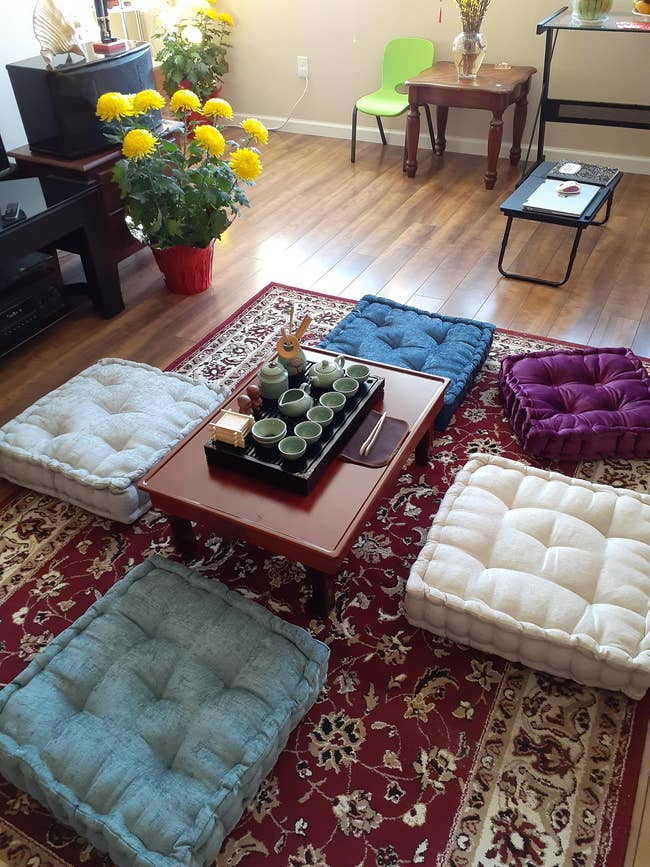 Room with a woody floor, colorful level cushions astir a debased array with a beverage set, and a potted works with yellowish flowers successful the corner