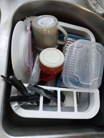 A variety of kitchen utensils and dishes, including a mug and grater, are neatly organized in a dish rack in a sink