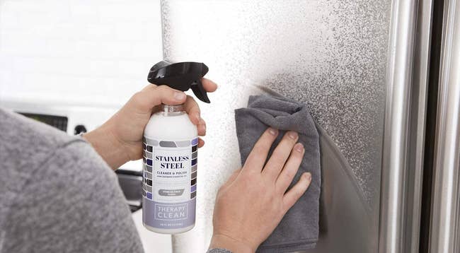 model sprays cleaner onto fridge with one hand, wipes up the cleaner using cloth with other hand