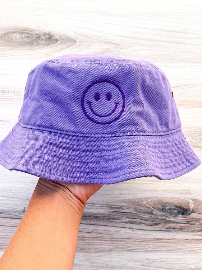 model holding a light purple bucket hat with a dark purple smiley face on it