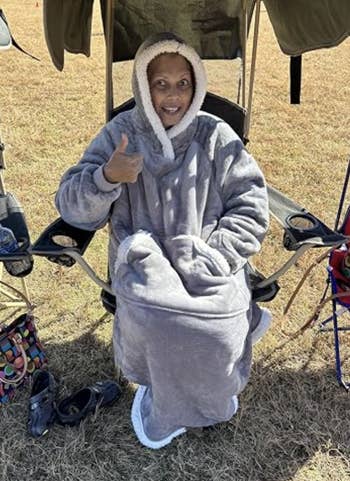 Person sitting successful  a camping seat  warmly wrapped successful  a plush, hooded blanket, giving a thumbs-up. Camping cogwheel  and shoes disposable   nearby
