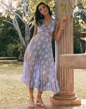 model in a mid-length flowy dress with a nature-inspired print, standing by a column, touching a vine. She wears strappy sandals