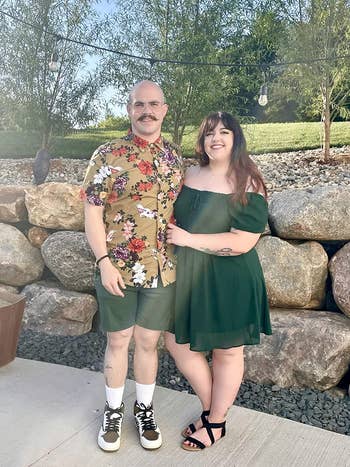A mates stands outdoors. The antheral wears a floral garment and shorts; the pistillate wears an off-shoulder dress. Both are smiling and embracing