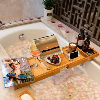 the tray on a bathtub holding an ipad, magazine, candle, snacks, phone, drink, and beauty products 