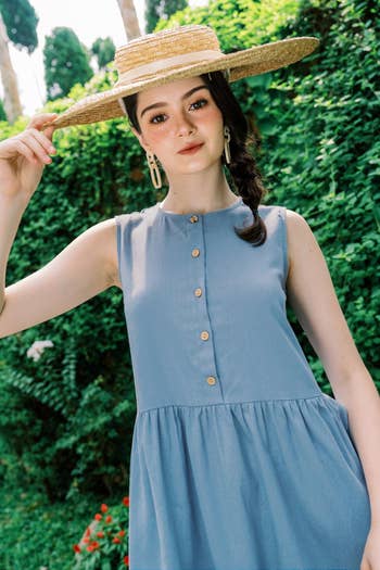 A woman wearing a sleeveless button-up dress, holding the brim of a wide-brim hat. She is standing outdoors in a garden