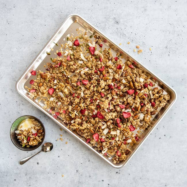 Tray of granola with dried effect and seeds adjacent to a tiny vessel of the aforesaid granola served with yogurt and a spoon