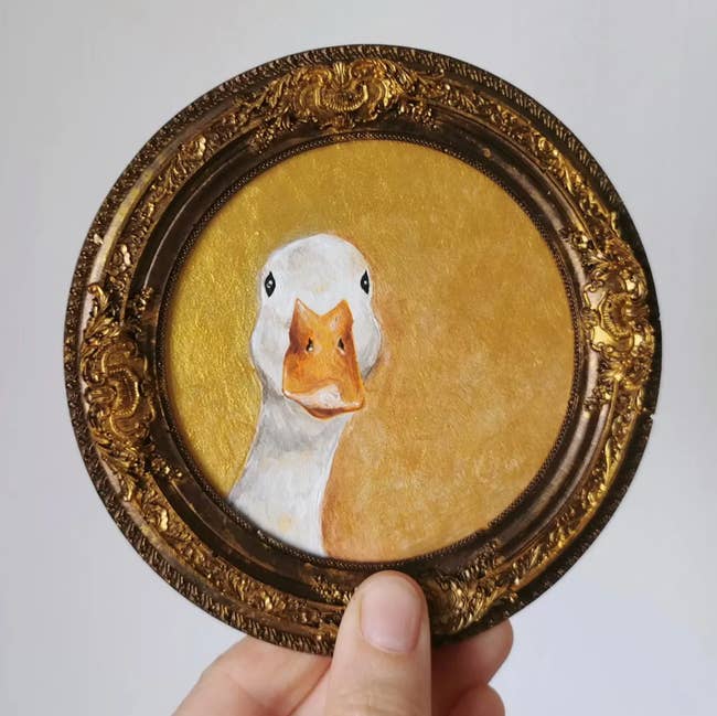A hand holds a small ornate frame with a painted portrait of a goose on a golden background