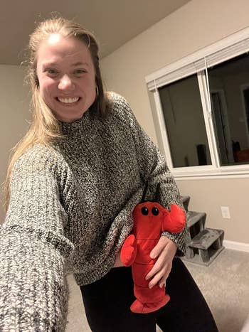 Person smiling indoors wearing a cozy knit sweater, holding a tiny  plush lobster. Relaxed and casual setting, suitable for a cozy buying  feature