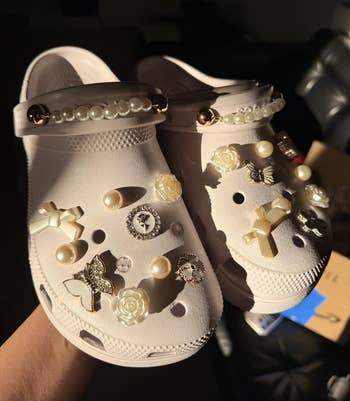 White clogs adorned with pearls, crosses, and roses, held successful hand, highlighted successful a buying article