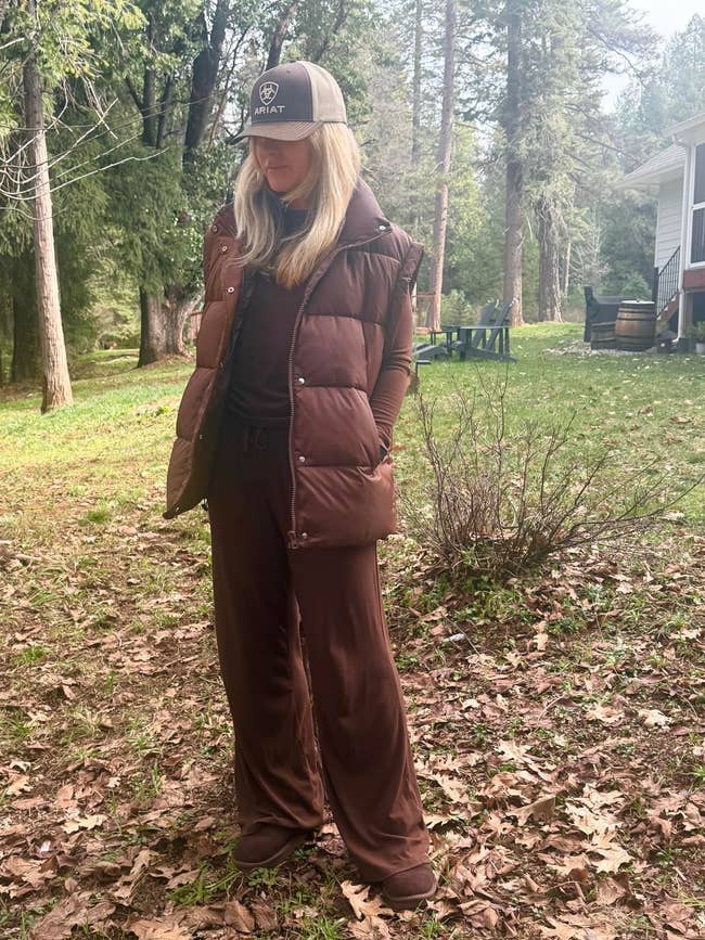 Person outdoors wearing a puffer vest, wide-leg pants, long-sleeve top, and shot   cap, lasting  connected  a leafy crushed  adjacent   a location   and trees