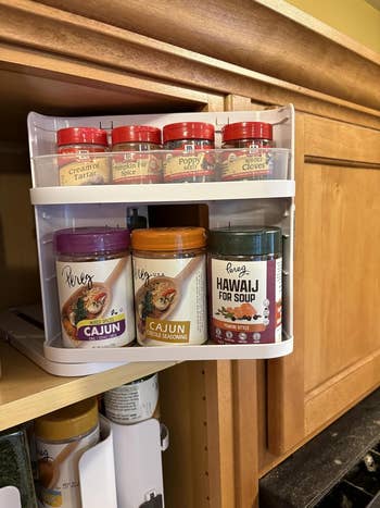 Kitchen rack with assorted  spice jars, including Cajun seasoning, hawajj for soup, and cloves, is mounted connected  a woody  cabinet
