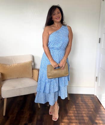 reviewer in a one-shoulder, tiered blue dress holding a woven clutch, standing next to a chair with pillows