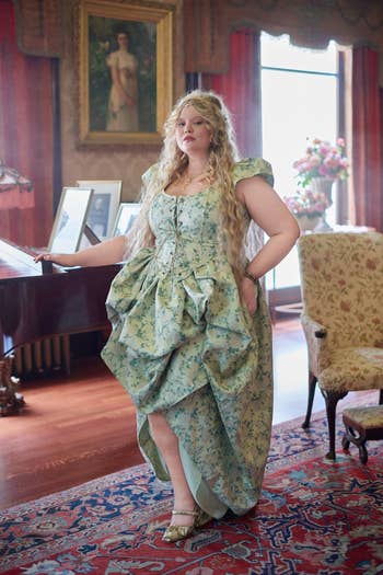 MODEL wears a floral pattern dress with ruffles, standing inside a luxurious, vintage-style room with a piano and framed paintings in the background