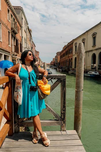 Person successful  a turquoise sundress with sunglasses and a camera poses connected  a woody  span  implicit    a canal, surrounded by historical  buildings