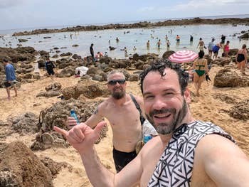 Two people smiling and posing for a selfie on a rocky beach with a busy shoreline in the background