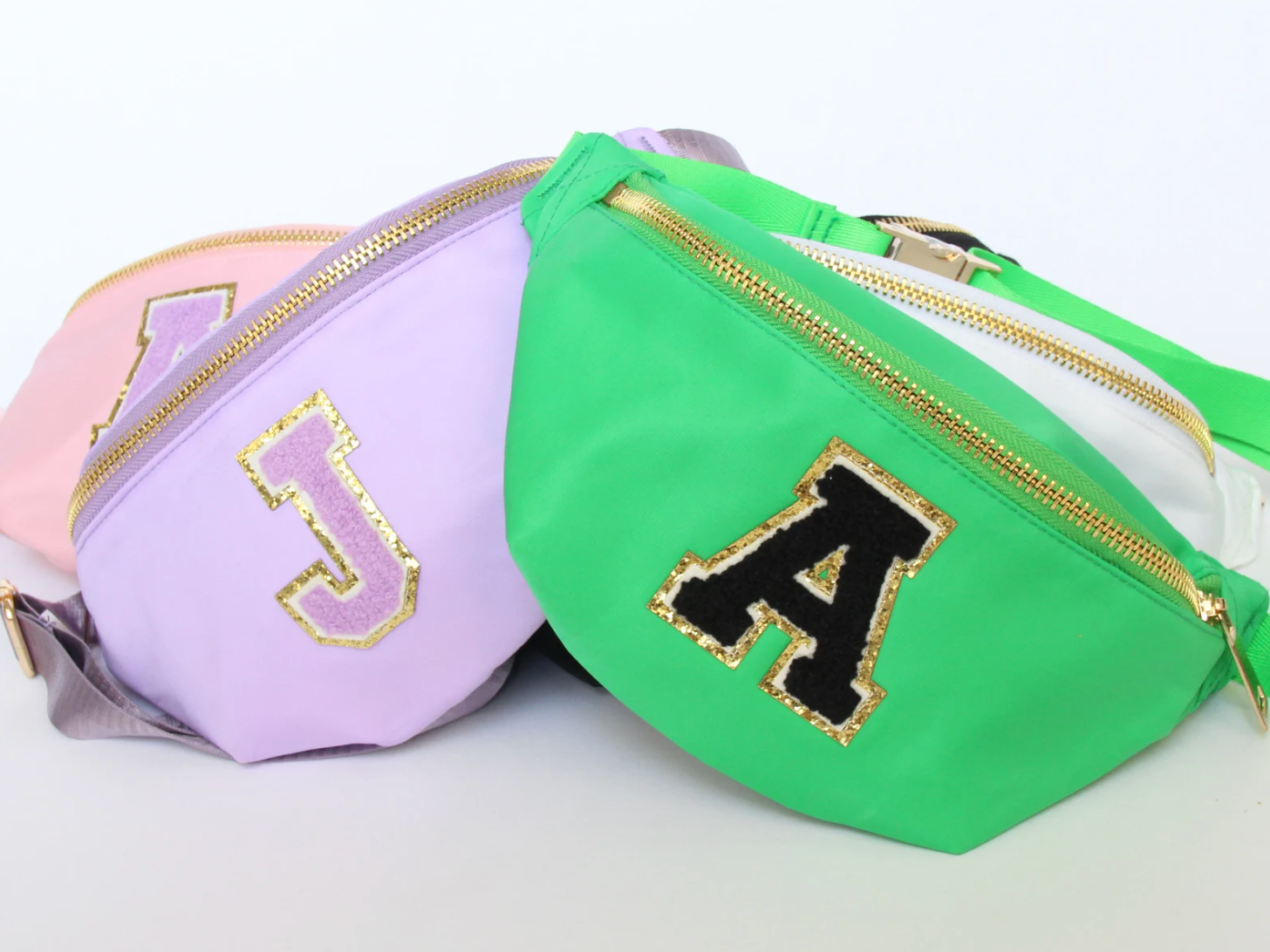 Green, purple, and pink fanny packs with patch letters A and J and gold sequins outlining them