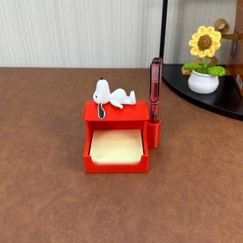 Snoopy-themed pen holder with a red roof, holding sticky notes. A small Snoopy figure rests on top