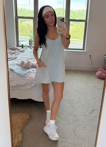 Person successful a casual sleeveless dress, headband, and sneakers taking a reflector selfie successful a chamber with a neatly made furniture successful the background
