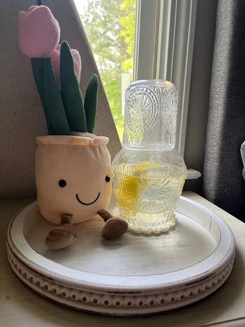 Smiling, plush flowerpot toy next to an ornate glass filled with lemonade on a white tray