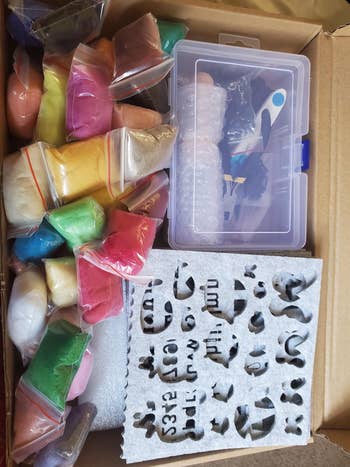 A container  containing assorted  colored sands successful  bags, a stencil with shapes, and coating  tools, suggesting a DIY creation  kit for crafting projects