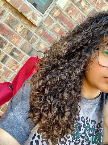 Person with curly hair wearing glasses and a graphic tee, partial face visible, sitting against a brick wall