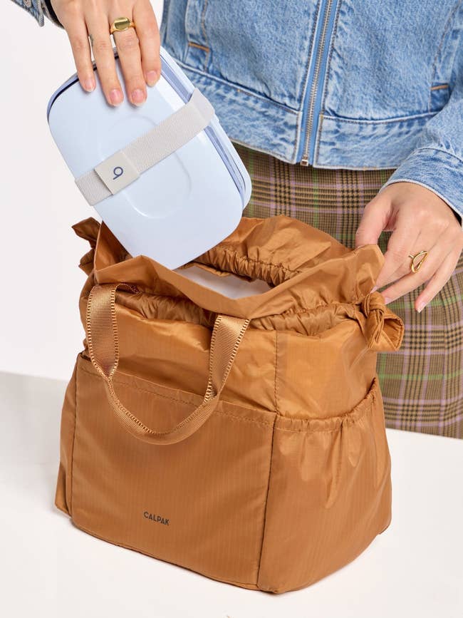 Person placing a bluish lunchbox into a brownish CALPAK bag, wearing a denim overgarment and plaid pants