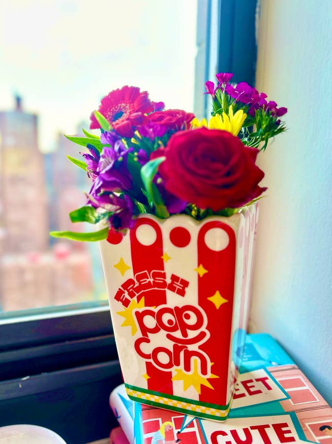 A bouquet of assorted flowers arranged in a popcorn-themed vase on a windowsill