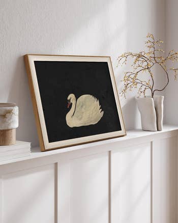 Framed painting of a swan on a shelf beside a vase with twigs, in a home setting