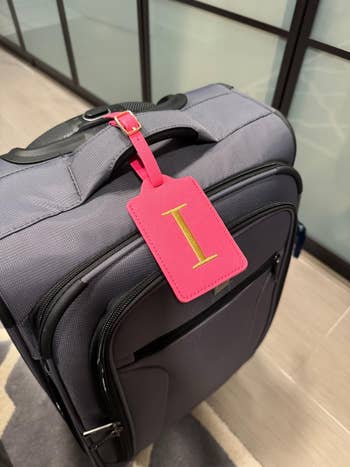 A grey suitcase with a pinkish luggage tag with the missive I