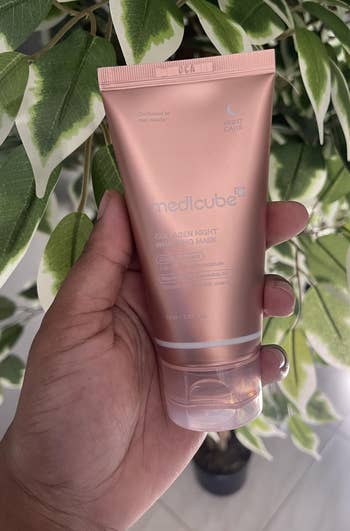 Person holding Medicube Collagen Night Wrapping Mask successful beforehand of a leafy plant. Product is simply a skincare item