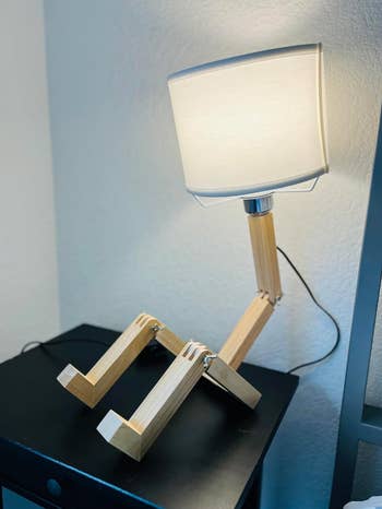 Wooden lamp with jointed legs and a boxy lampshade, resembling a seated quality  figure, connected  a achromatic  table