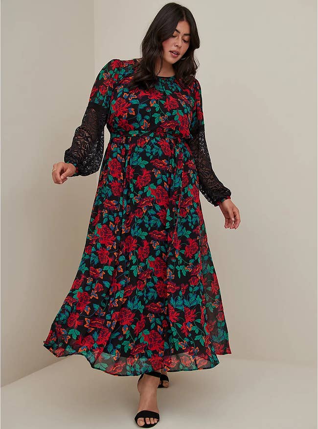 Model wearing maxi crew neckline dress with red floral pattern and black lace longsleeves paired with black open-toe heels on a white background
