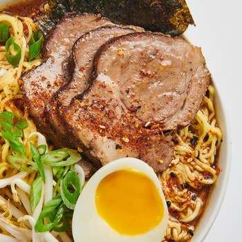 A vessel  of ramen with sliced pork, scallions, legume  sprouts, nori, and a halved soft-boiled egg, showcasing a delicious, savory repast  option