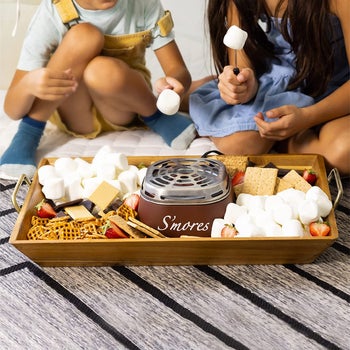 two models using s'mores maker surrounded by marshmallows, graham crackers, and other food items