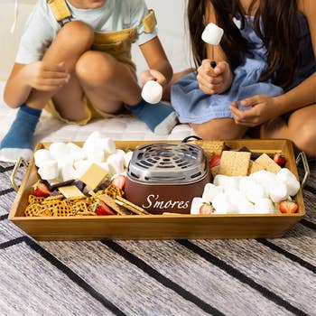 two models using s'mores maker surrounded by marshmallows, graham crackers, and other food items