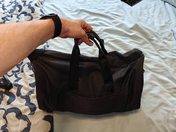 A hand holds a black duffel bag with top handles over a bed with a patterned blanket