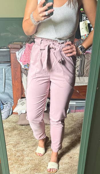 Person in sleeveless top, high-waisted belted pants, and sandals taking a mirror selfie in a cozy room